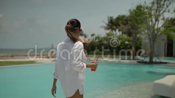优雅的年轻女士在酒店游泳池附近享受美好的一天穿着白色衬衫拿着一杯带吸管的红色果汁视频的预览图