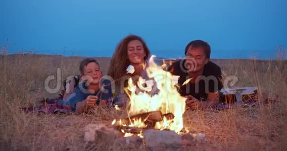 儿子和他的父母用棍子烤棉花糖晚上家人在海滩露营一个家庭视频的预览图
