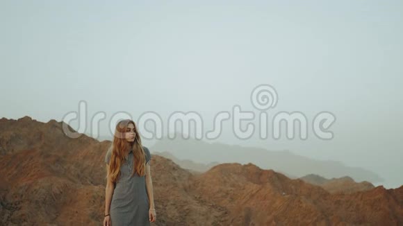 年轻的金发女人穿着长裙走在山顶上带日落的山景慢动作全高清视频的预览图