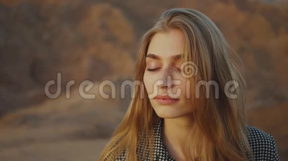 美丽的金发女人的肖像闭着眼睛看日落快乐的年轻女子站在沙漠中日落视频的预览图