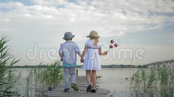 快乐可爱的孩子男孩和女孩带着玩具风车手牵手地走在绿色芦苇之间的河边的木桥上视频的预览图