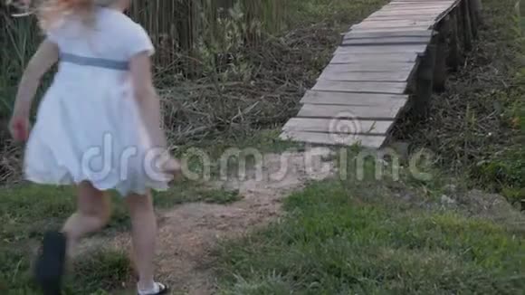 穿着白色连衣裙的活泼可爱的小女孩沿着大自然中的木桥在绿色的植被中奔跑视频的预览图