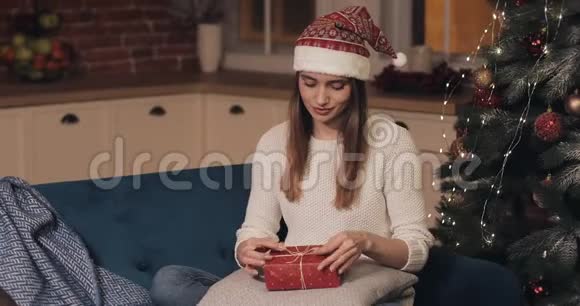 戴着圣诞帽的年轻女性在舒适的圣诞背景下坐在沙发上手握红礼视频的预览图
