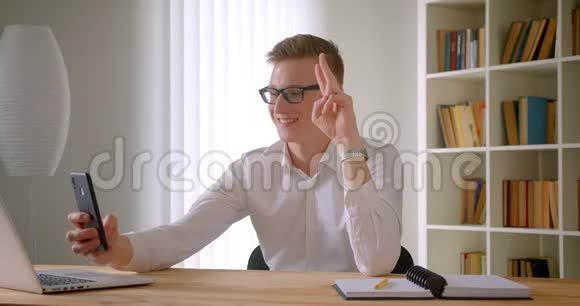 年轻英俊的白种人商人戴着眼镜在手机上自拍的特写肖像露出两根手指视频的预览图