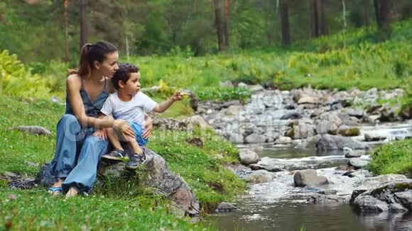年轻的母亲和小儿子在山溪边聊天坐着视频的预览图