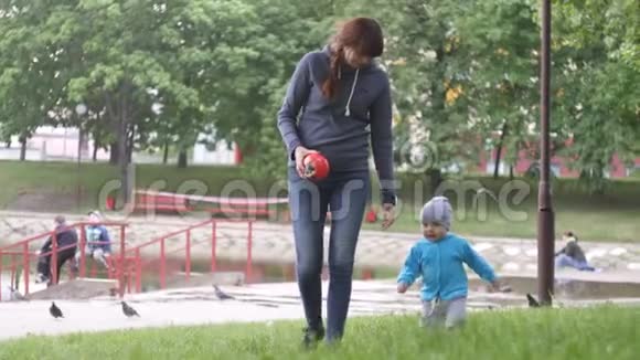 在一个夏天的阴天里带着她的小儿子的年轻女子在城市公园里散步池塘和树在那里视频的预览图