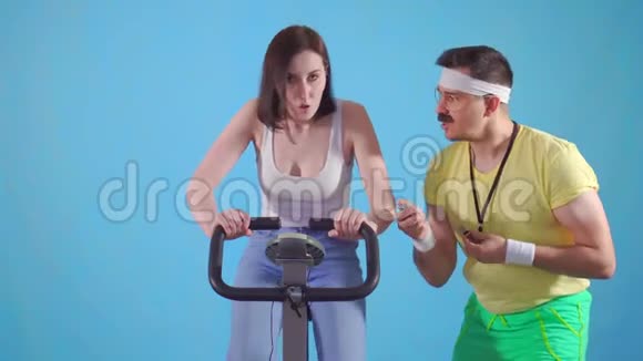 有趣的男子教练80带胡子训练一个年轻女子骑着蓝色背景的运动自行车视频的预览图