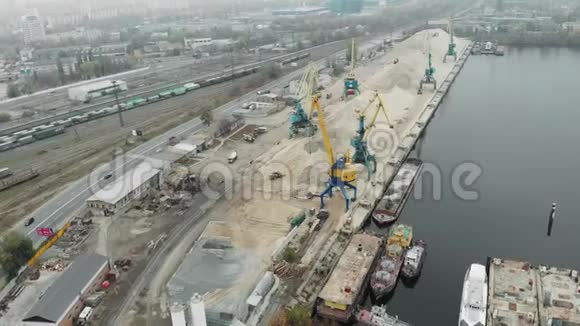 在烟雾弥漫的工业肮脏城市中港口起重机从铁驳船和河堤上提取沙子的鸟瞰图视频的预览图