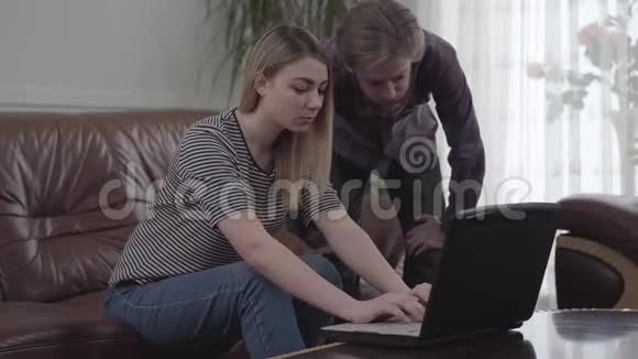 漂亮的女人坐在皮革沙发上胡须男站在她身边拿着笔记本电脑工作小女孩带着视频的预览图
