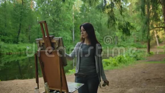 美丽的女艺术家在池塘附近的公园里画一幅风景画视频的预览图