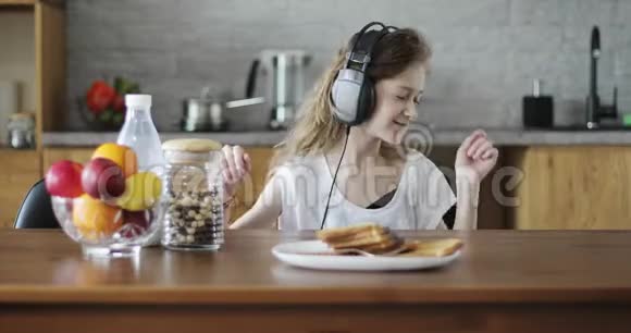 快乐的女学生带着耳机坐在厨房的桌子旁跳舞视频的预览图
