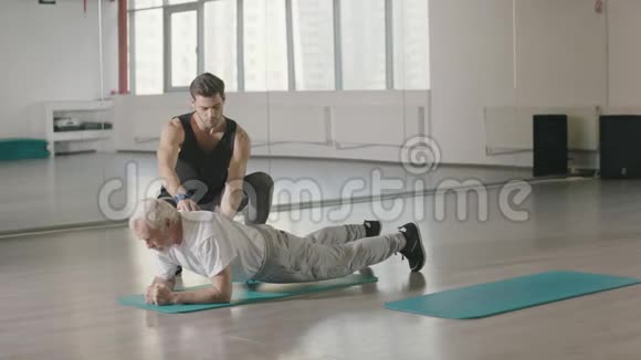 老年人与体育教练一起在健身俱乐部训练木板锻炼视频的预览图