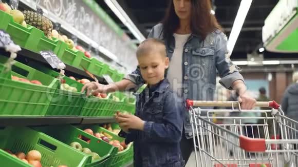 可爱的金发男孩正和他的母亲一起买水果他正从塑料盒子里拿出苹果放在手推车里视频的预览图