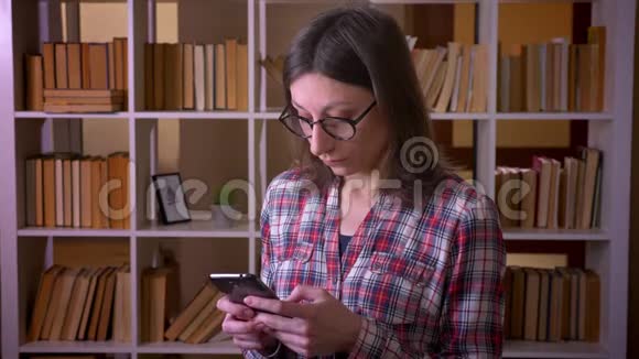 在室内使用手机和看着图书馆里的摄像机用眼镜拍摄年轻迷人的女学生的特写镜头视频的预览图