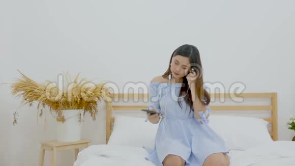 4K美丽的亚洲女人坐在床上用手机上的耳机听音乐跳舞视频的预览图