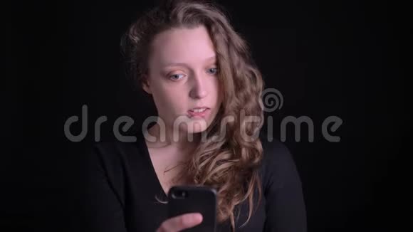 年轻的卷发女孩在黑色背景下使用智能手机在电视上愉快地交谈的肖像视频的预览图