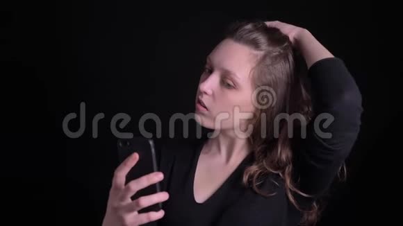 年轻卷发女孩在黑色背景下愉快地使用智能手机制作自拍照片的肖像视频的预览图