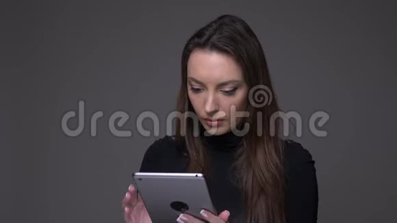 漂亮的黑发女人将绿色的平板电脑屏幕显示在相机中以推荐隔离在灰色上的应用程序视频的预览图