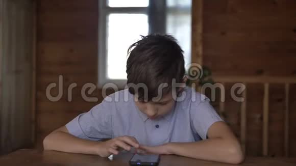 有趣的男孩坐在家里的桌子上用电话玩游戏视频的预览图