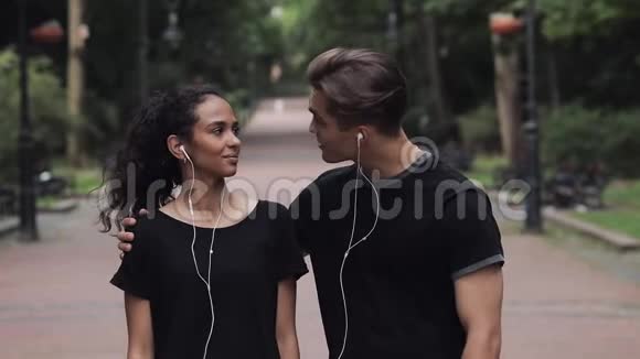 迷人的音乐女孩和戴着耳机说话的年轻人在公园慢悠悠地拥抱她视频的预览图