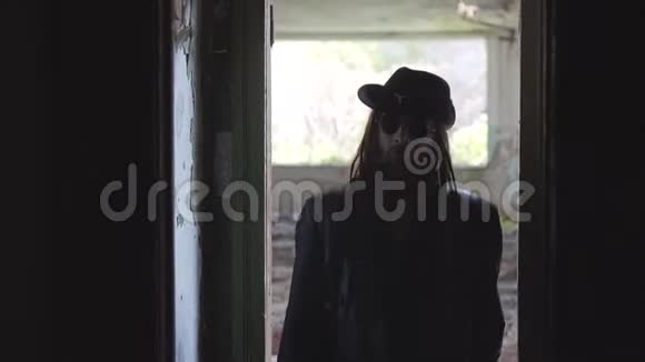 一个穿着黑色雨衣和帽子的酷男人走进一座废弃建筑的黑暗走廊拿着一把刀在移动视频的预览图