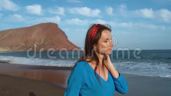 戴着太阳镜和蓝色连衣裙的女人沿着黑色的火山海滩散步慢动作视频的预览图