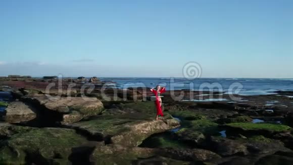 在黑色沙滩附近的岩石上展示美丽性感的比基尼女郎和红色的整体造型视频的预览图