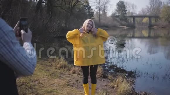 年轻的金发女人在森林和湖边摆姿势男人用手机给她拍照视频的预览图
