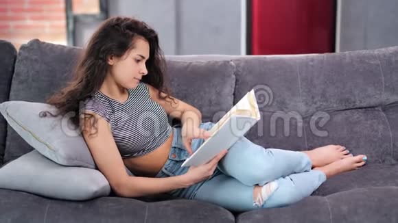 专注的年轻家庭女性躺在舒适的沙发上看书全镜头视频的预览图