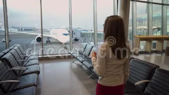 一个年轻漂亮的女孩走近机场的全景窗口透过那里可以看到飞机视频的预览图