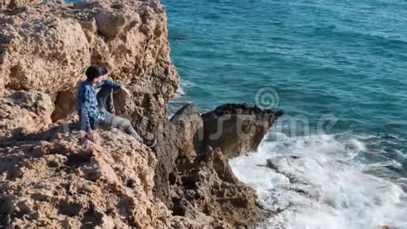 穿着蓝色衬衫和帽子坐在岩石上看着大海的年轻人坐在悬崖边上的人男性游客在岩石上休息视频的预览图