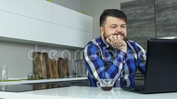一个大胡子的家伙盯着笔记本电脑超重的人喜欢他的咖啡早上坐在厨房里厚男性视频的预览图