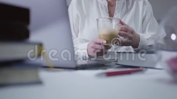 无法辨认的白种人成熟妇女喝卡布西诺和使用笔记本电脑资深退休人士在家上网视频的预览图