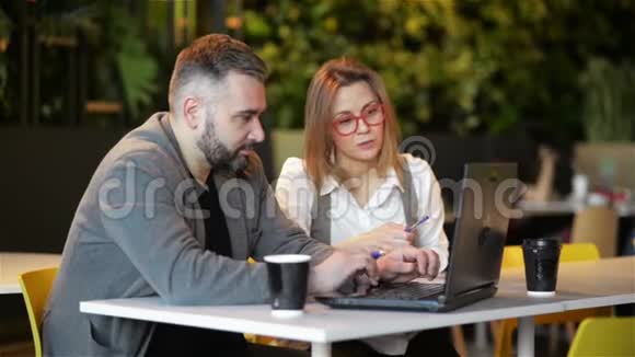 两位年轻的专业人士坐在办公桌前看着笔记本电脑屏幕一起讨论项目女孩在喝酒视频的预览图