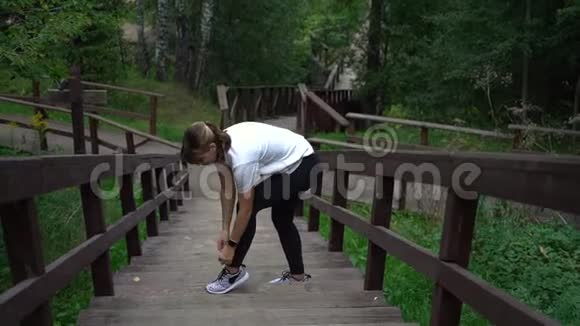 漂亮的黑发高级适合女性系鞋带在运动鞋在木楼梯在公园穿运动服装领带的苗条女人视频的预览图