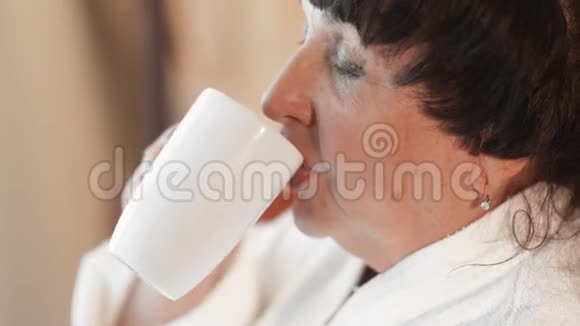 一位穿着白色特里长袍的白人高级妇女早晨在一扇大的全景窗户附近喝咖啡慢动作视频的预览图