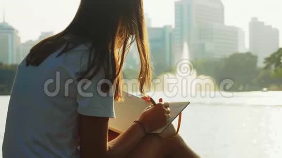 女人正在她的记事本上写着她坐在池塘附近的城市公园里有喷泉视频的预览图