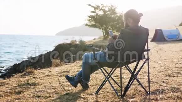 年轻人坐在文件夹椅上在湖岸露营时使用智能手机视频的预览图