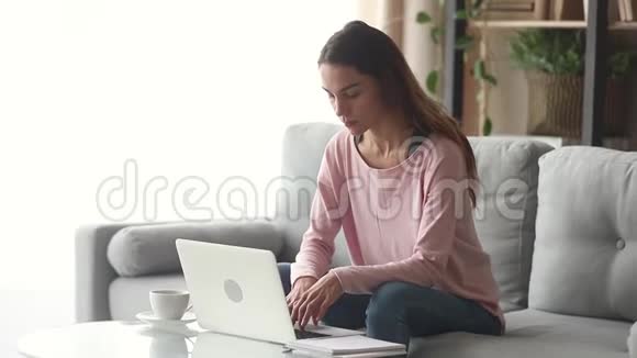 严肃快乐的年轻女人坐在沙发上用笔记本电脑视频的预览图