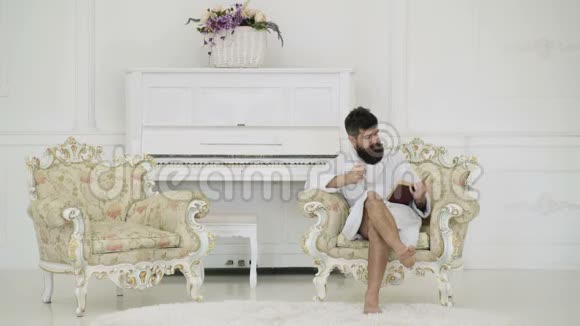 穿着浴袍的男人喝着咖啡坐在扶手椅上看着一本书背着一架白色的钢琴有胡子的男人视频的预览图