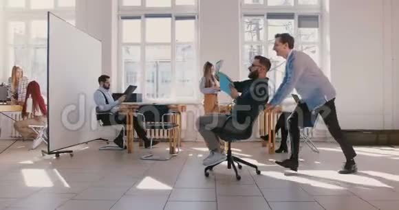 多利拍摄了疯狂有趣的商人骑着办公椅沿着办公大厅庆祝和扔纸在空中视频的预览图