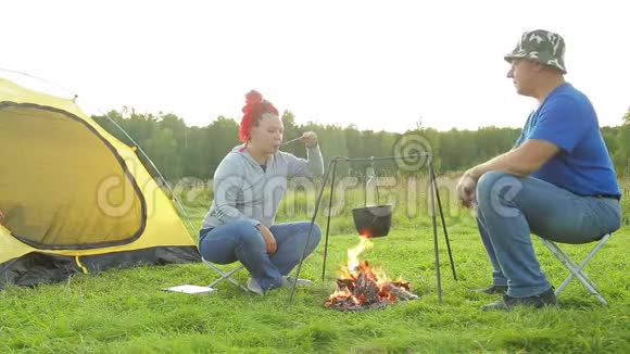 在一个阳光明媚的夜晚一对男女在靠近火的帐篷旁用锅做饭视频的预览图