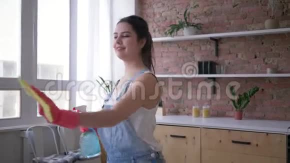 快乐的家庭主妇穿着牛仔工作服戴着手套在清洁时手里拿着清洁剂喷雾剂和清洁剂跳舞视频的预览图