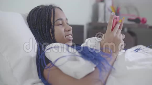 非洲裔美国女孩的肖像可怕的躺在白色的床上使用智能手机年轻女子上网视频的预览图