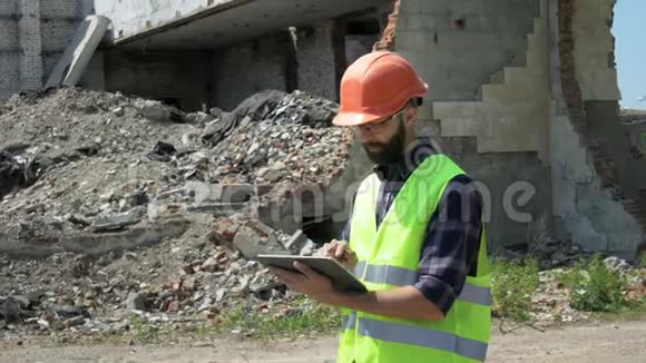 戴着头盔和反光背心的长胡子建筑工人在被摧毁的建筑背景上使用平板电脑为准备视频的预览图