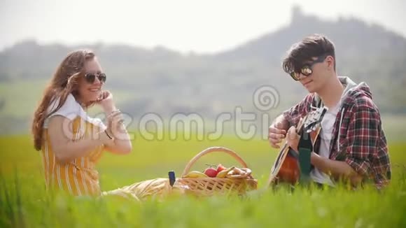 一对年轻夫妇坐在明亮的绿色草地上野餐一个男人弹吉他而他的幸福女友视频的预览图