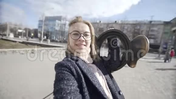 漂亮的微笑金发女人在时尚的外观自拍与雕塑形式的耳朵在街上时尚博客视频的预览图