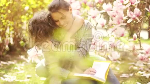 快乐的少年拥抱他美丽的年轻母亲春天的肖像背景上盛开的粉红色玉兰母亲视频的预览图