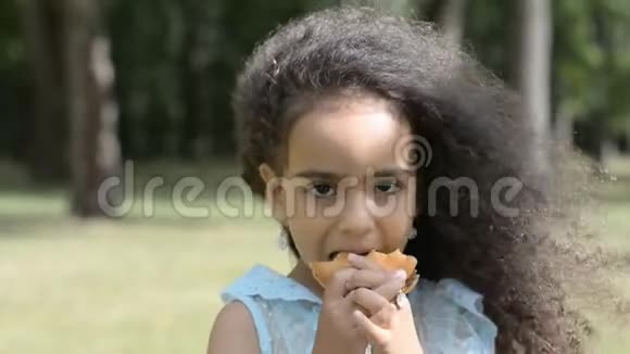 接近儿童脸非洲女孩肖像吃巨大汉堡与胃口在城市公园夏季或春季视频的预览图