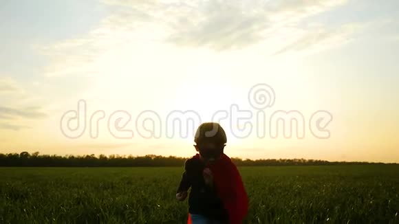 一个穿着超级英雄服装的快乐的孩子穿过草地在夕阳的背景下朝镜头跑去视频的预览图
